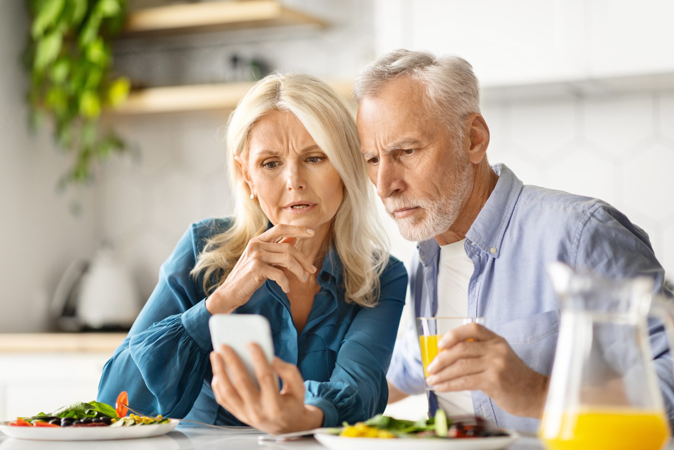 Concerned,Senior,Spouses,Reading,News,On,Smartphone,During,Breakfast,In
